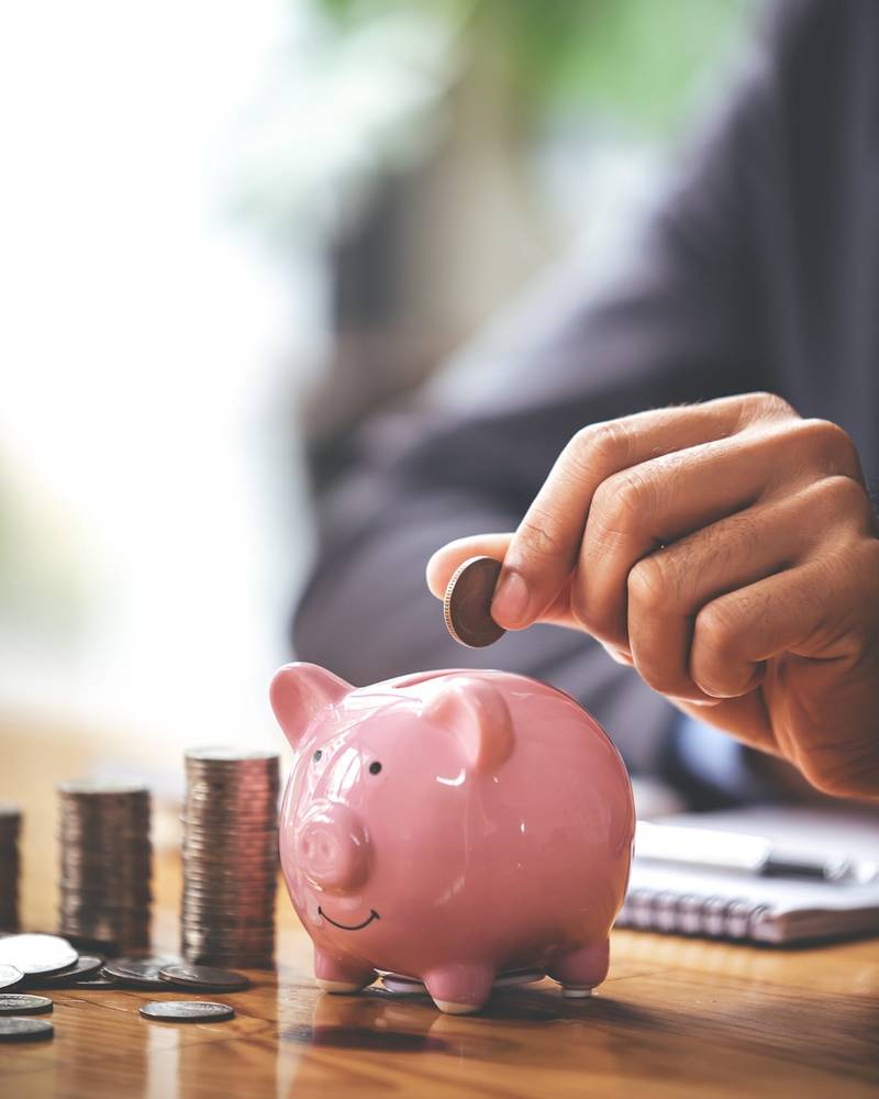 A hand dropping a coin into a piggy bank beside stacks of coins