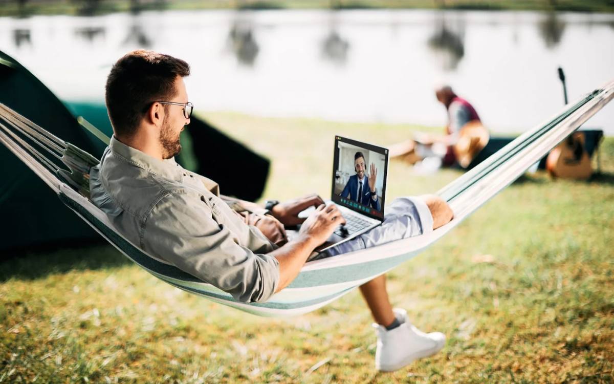 A man working on a laptop in a hammock outdoors, on a video call with someone waving from the screen