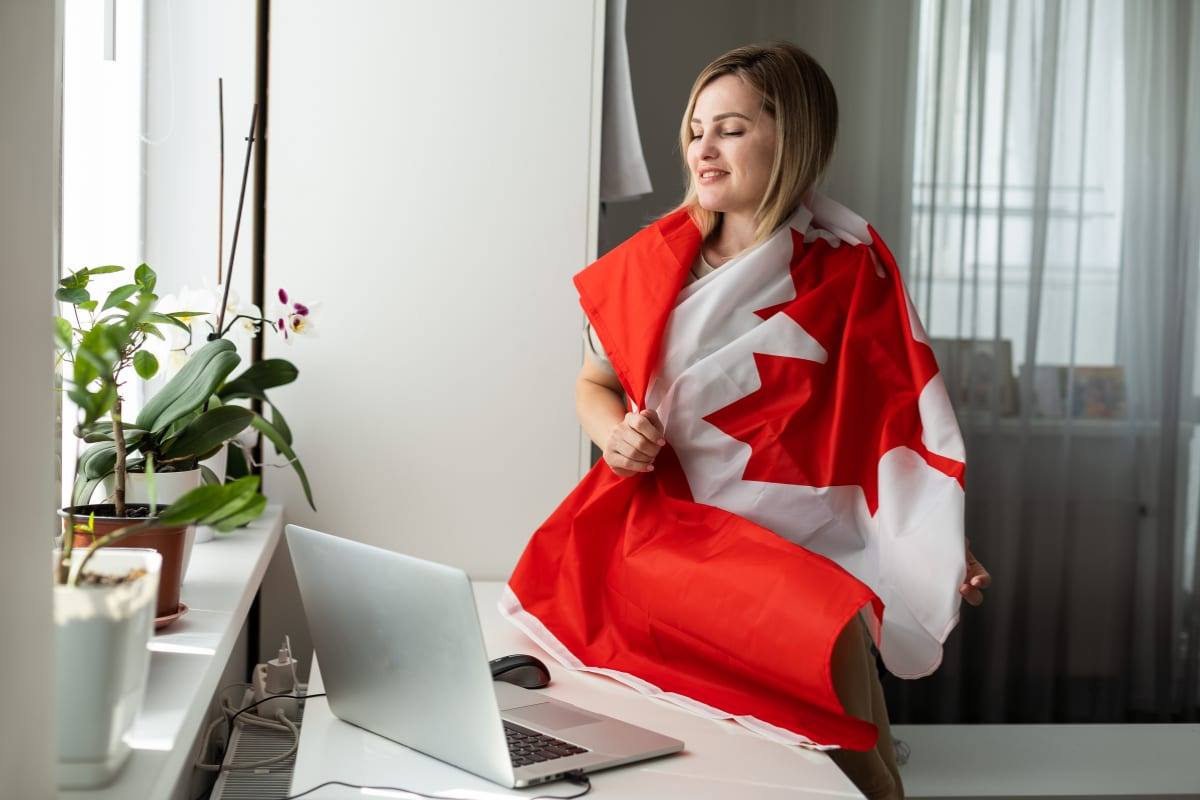 A woman draped in a Canadian flag standing by a home-office desk with a laptop and orchid