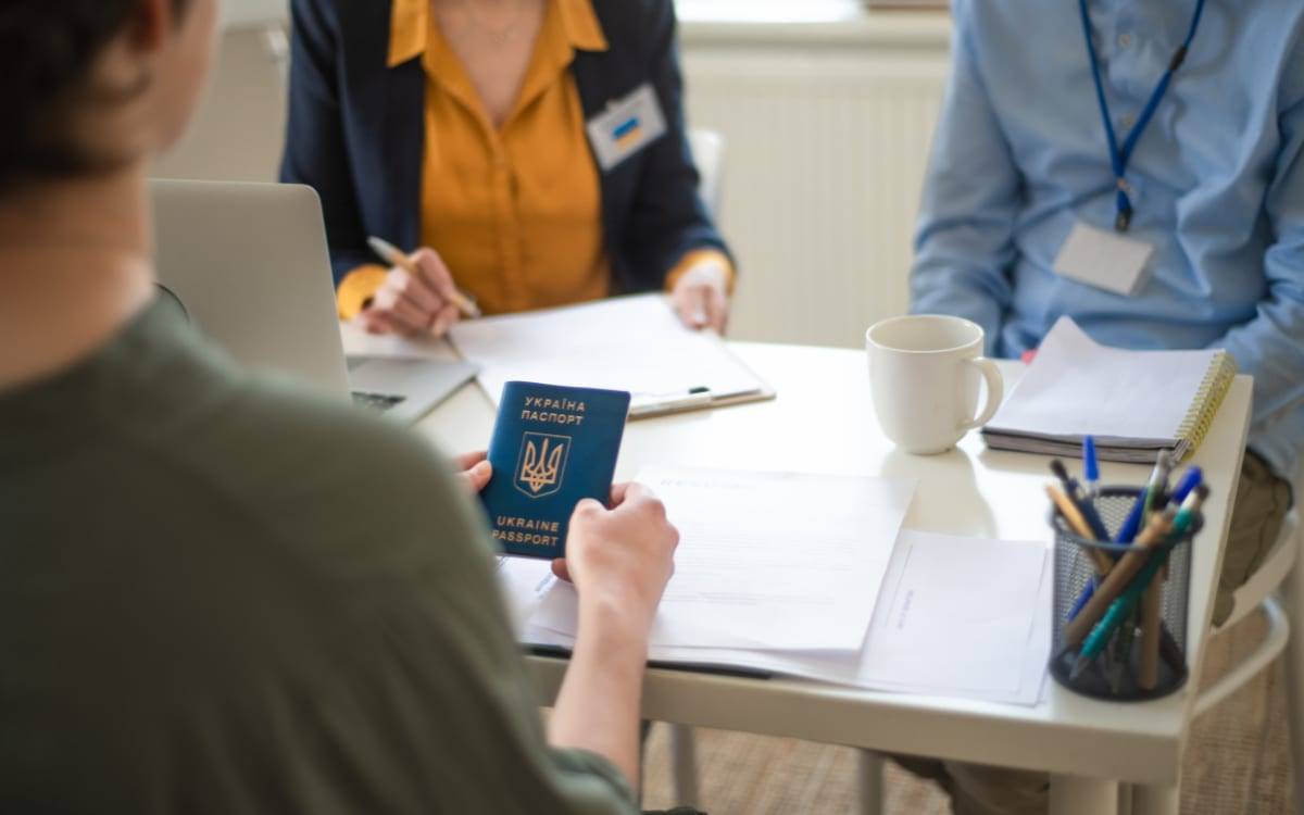 An applicant holding a passport across a desk from two officials during an interview