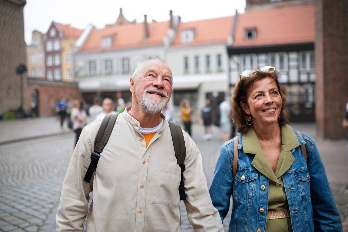 A smiling older couple with backpacks walking through a European old-town square