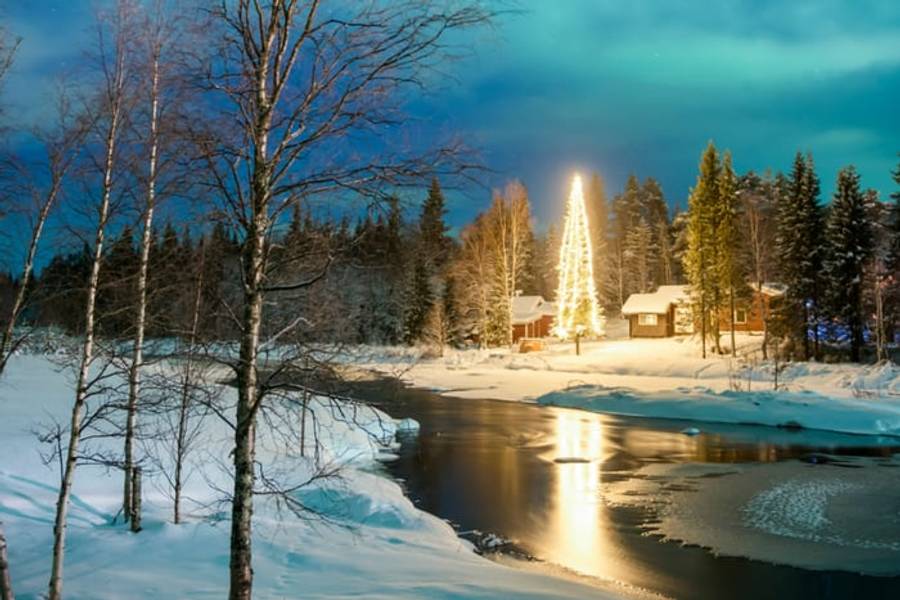 A lit Christmas tree beside a snowy riverside cabin in Lapland, Finland