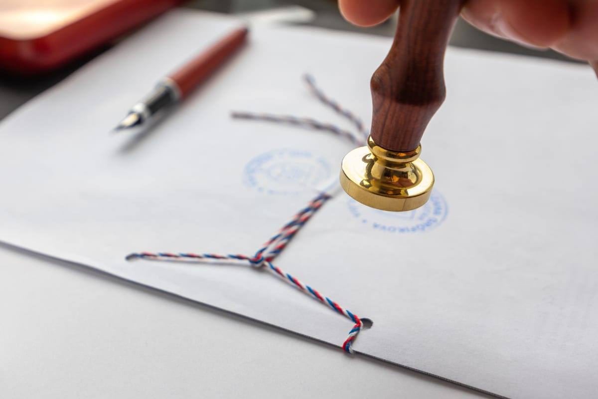 A hand pressing a brass seal onto an official document tied with tricolour string, beside a fountain pen