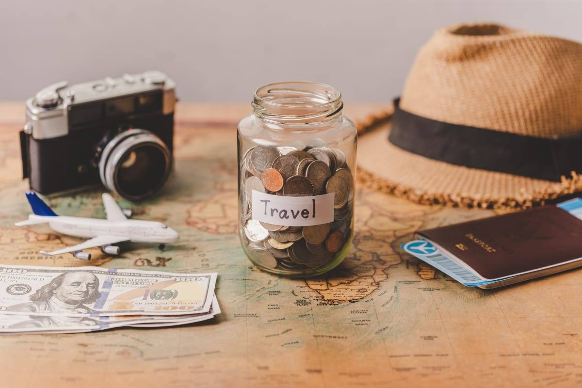 A jar of coins labelled Travel on a world map with a passport, US dollars, a camera and a sun hat