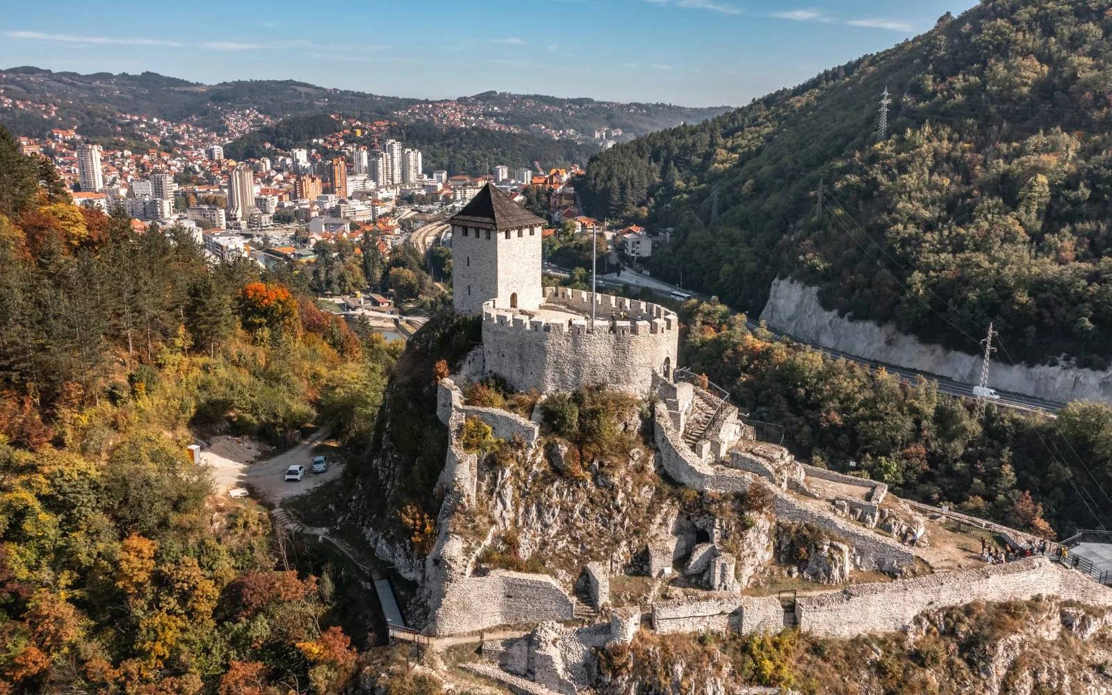 A medieval stone fortress on a rocky ridge above a town, with autumn hillsides and apartment blocks in the valley below