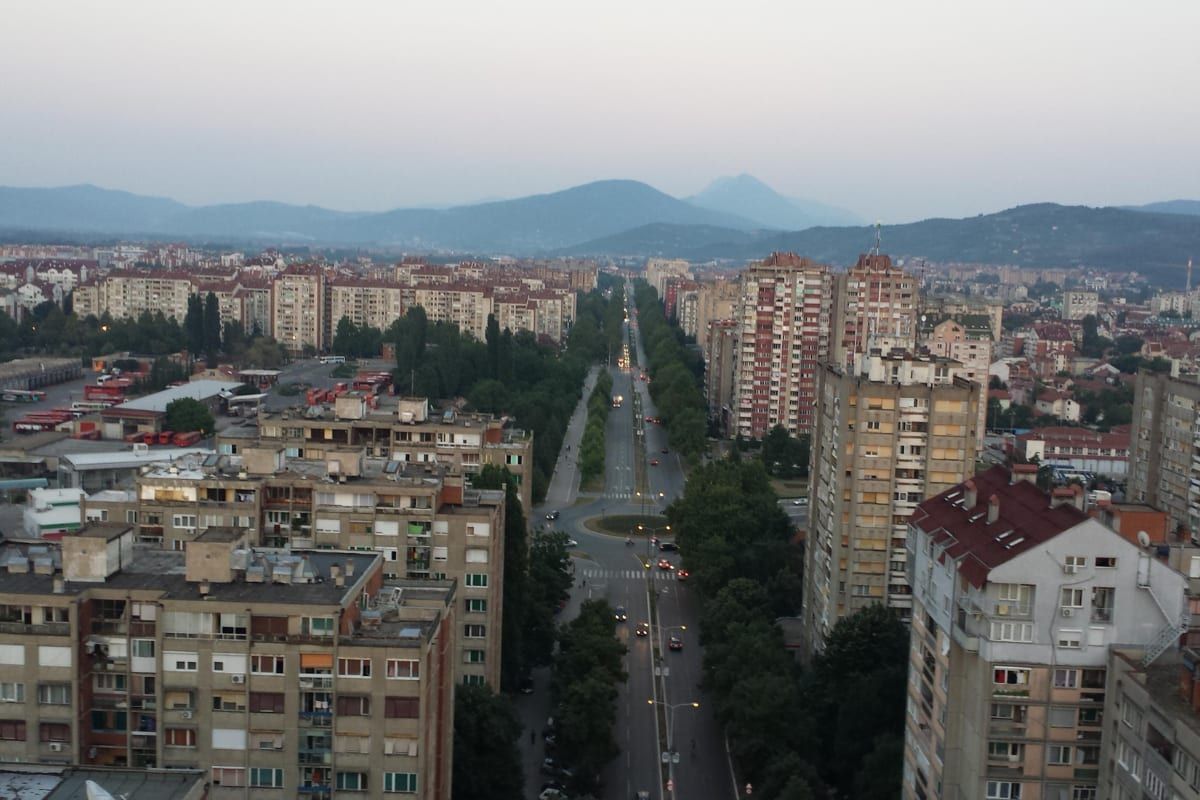 A long tree-lined boulevard flanked by tall apartment blocks in Niš at dusk, hills on the horizon.