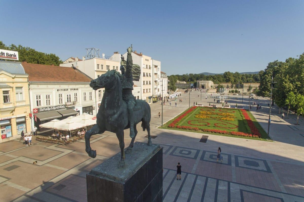 Niš's main square with the equestrian Liberators' Monument, café terraces, and a flower parterre, the fortress gate visible beyond.