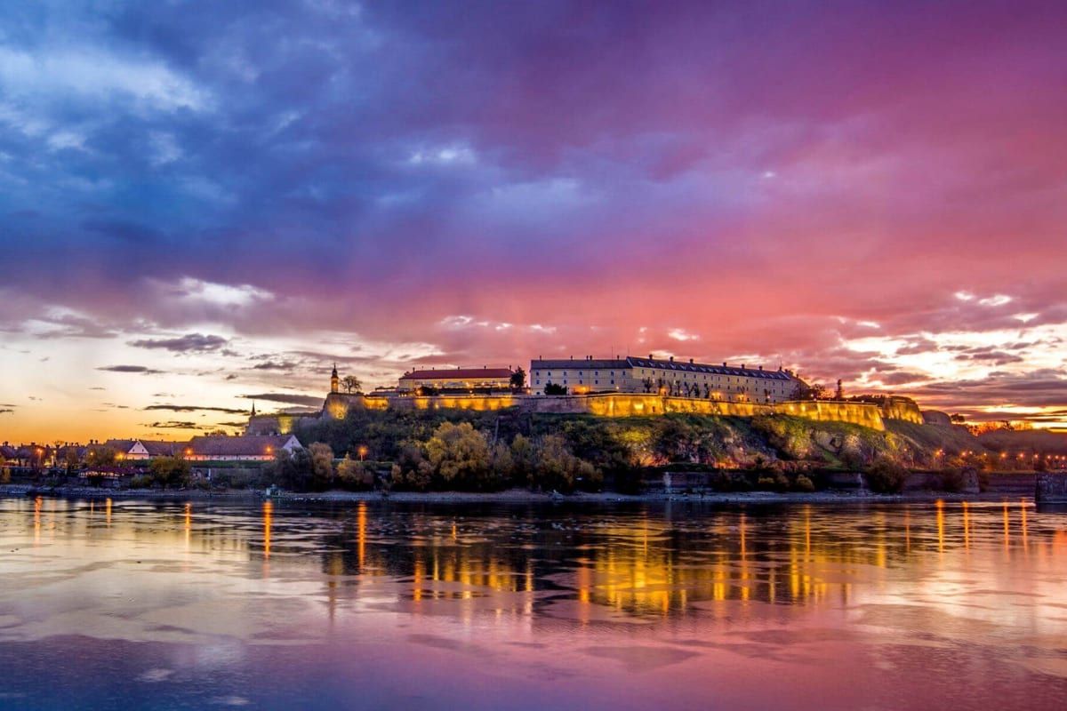 Petrovaradin Fortress lit above the Danube under a vivid sunset, across the river from central Novi Sad.