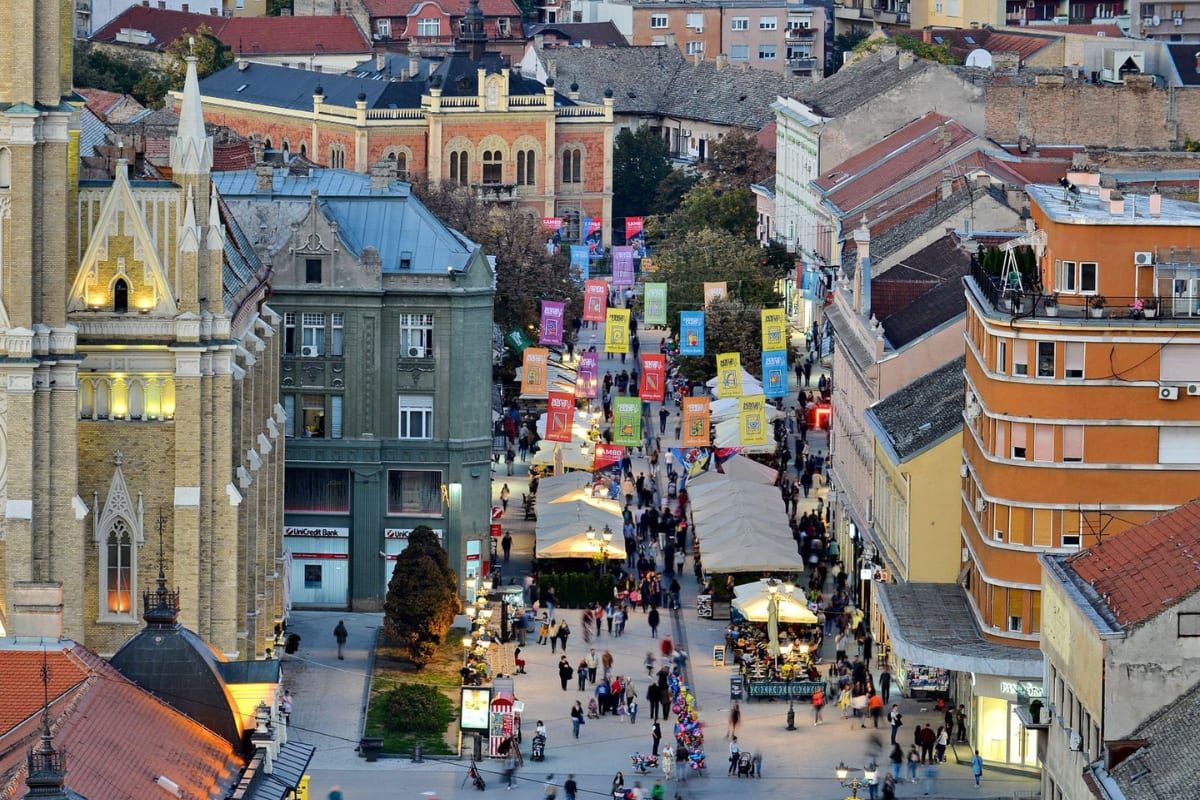 Aerial view down the pedestrian old town of Novi Sad at dusk, the neo-Gothic Name of Mary church spire on the left and market stalls lining the street.