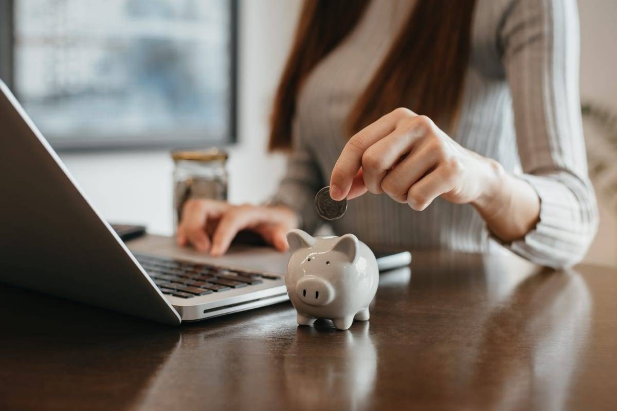 A woman dropping a coin into a piggy bank while working at a laptop