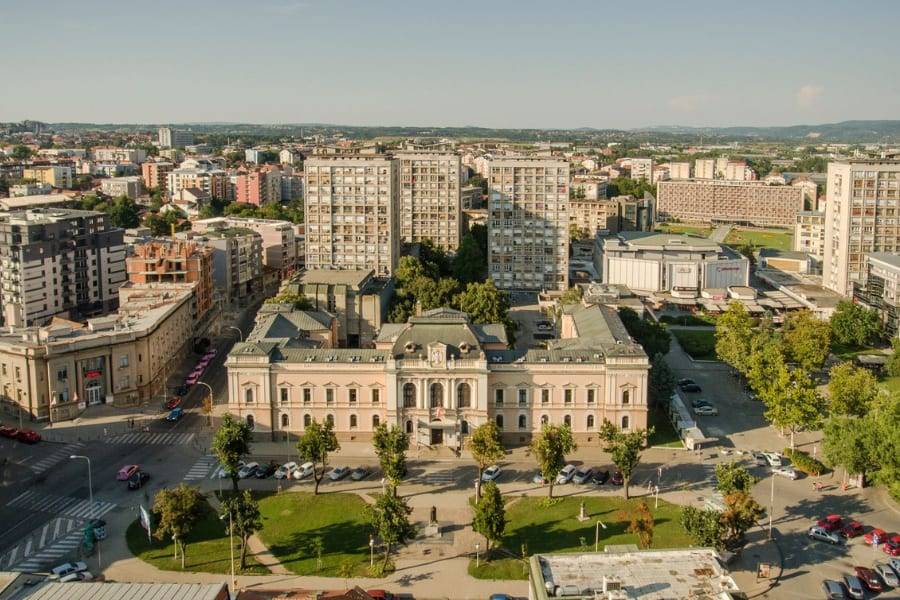 An aerial view of a Serbian town centre with a historic building, green square, and rooftops on a summer day