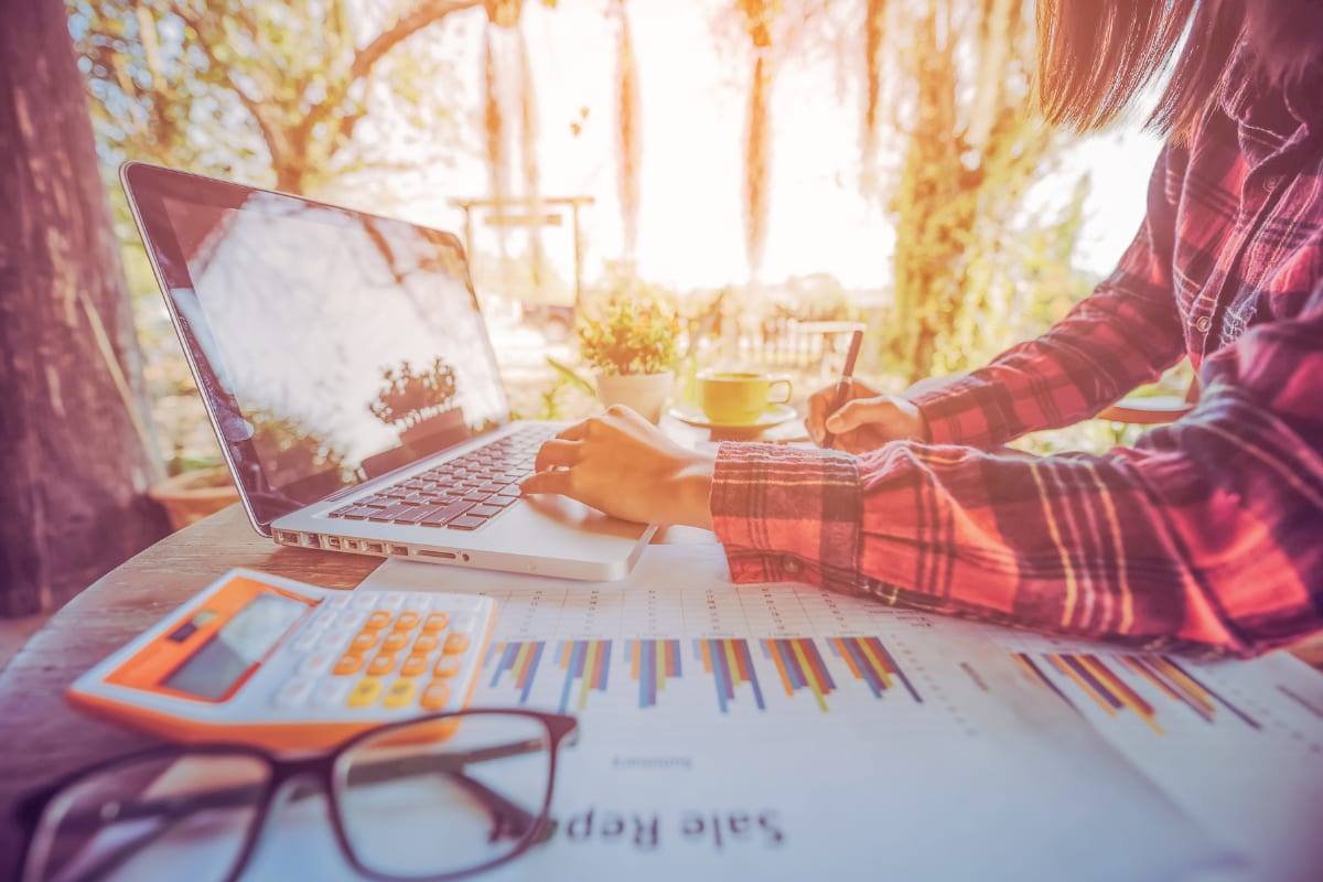 A freelancer working on a laptop outdoors with a calculator, glasses and a sales-report chart on the table
