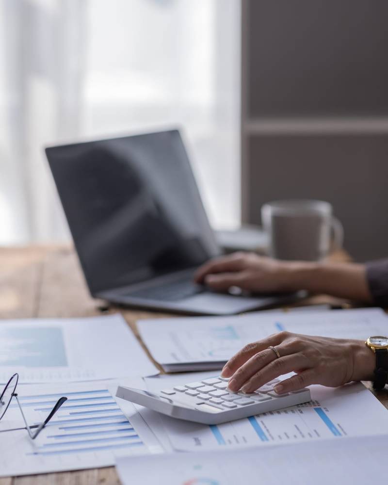 A hand using a calculator over payroll spreadsheets and charts, with a laptop behind