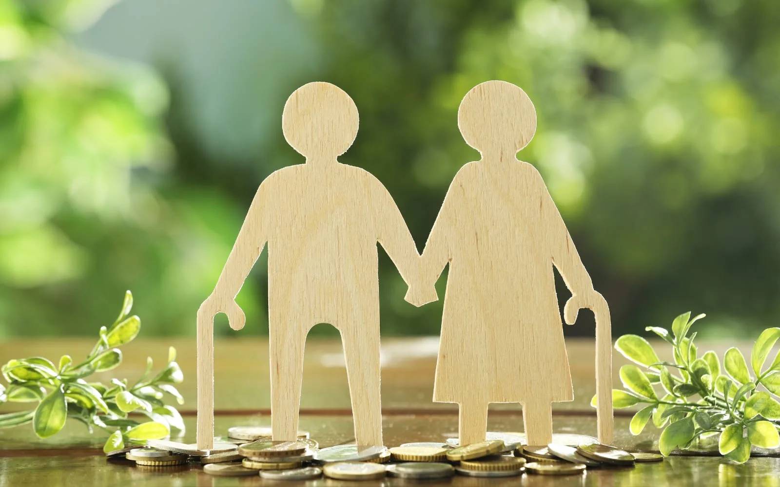 Two wooden cut-out figures of an older couple holding hands and leaning on canes, standing on a pile of coins