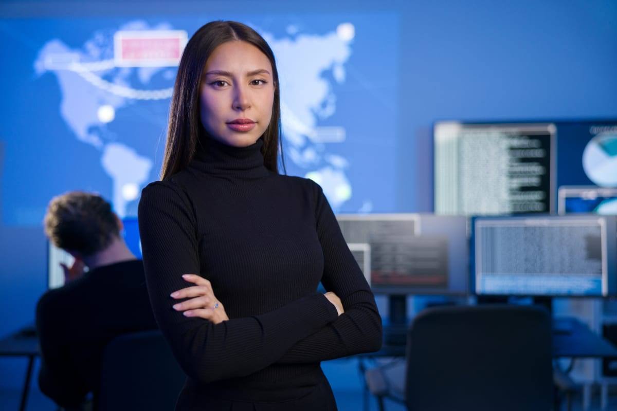 A confident woman in a black turtleneck standing in a blue-lit tech operations room with a world map on screen