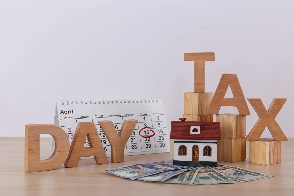 Wooden letters spelling TAX DAY beside a calendar with the 15th circled, a model house and US dollar notes