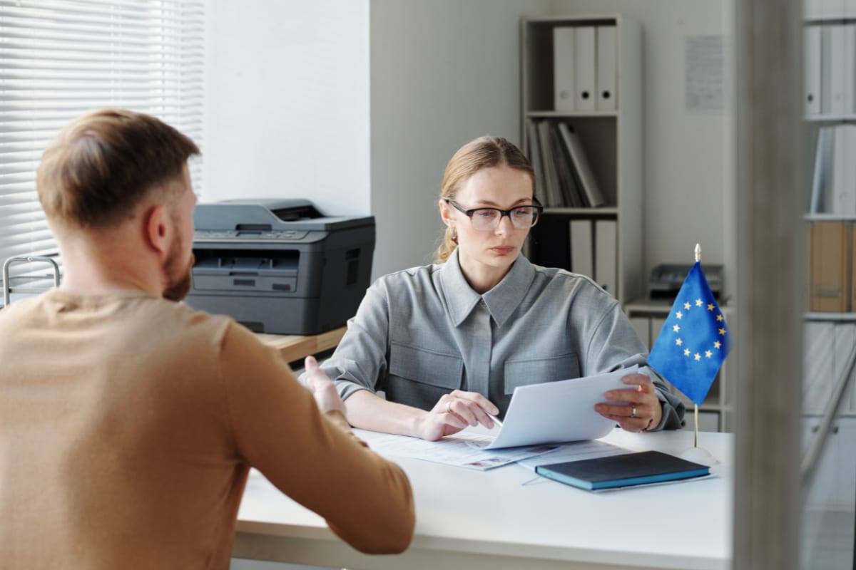 An applicant talking with an official reviewing documents at a desk beside a European Union flag
