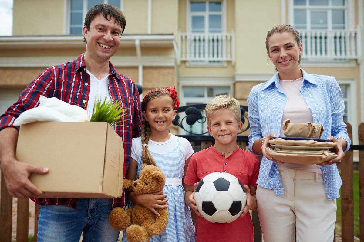 A smiling family of four carrying moving boxes and belongings in front of a house