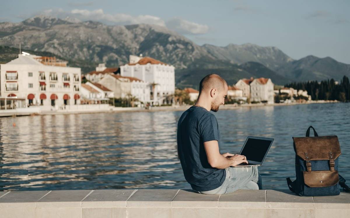 Un hombre trabaja con su laptop en un paseo marítimo, con mochila, un pueblo costero y montañas al fondo