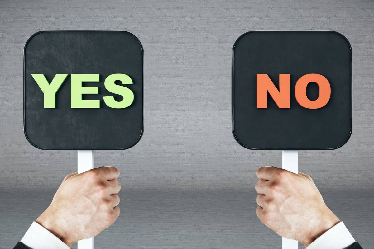 Two hands holding up a green YES sign and an orange NO sign against a grey wall